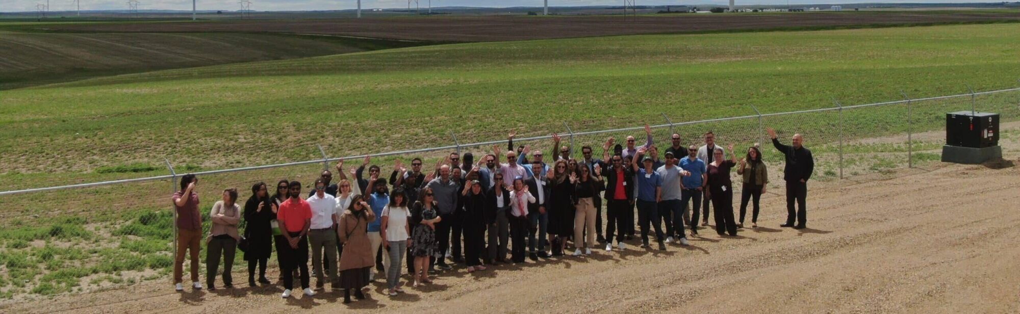 An above-landscape view of a group of approximately 60 people smiling and waving in front of four wind turbines in the distance behind them. It is a bright, windy day.