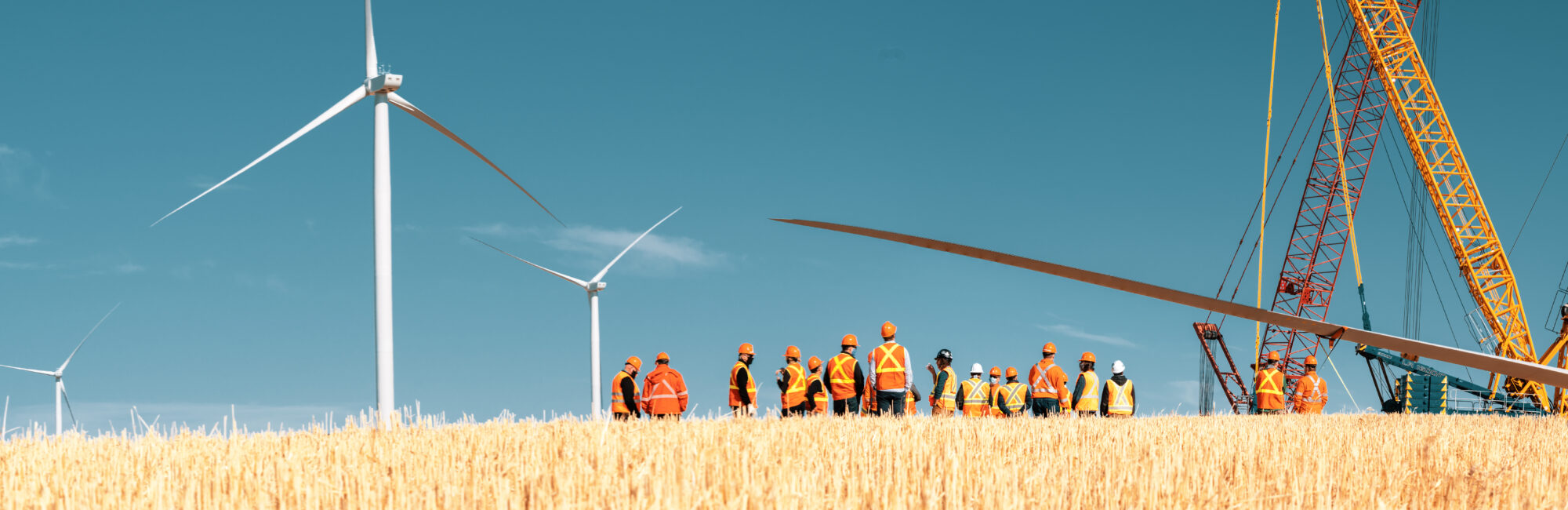 A row of construction workers in orange vests and hard hats stand looking at three running wind turbines.