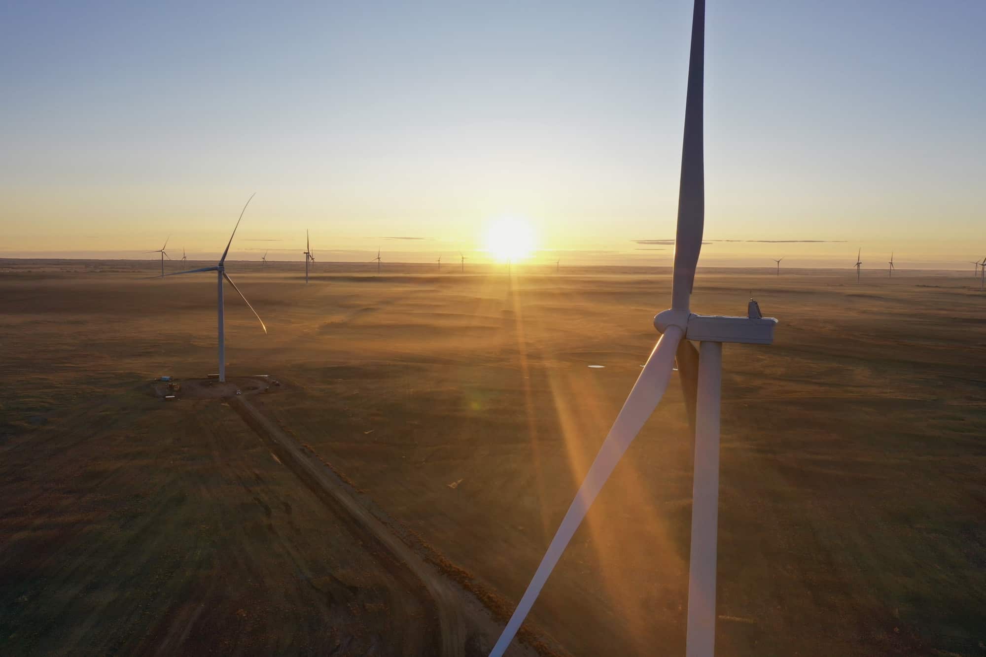 A landscape photo of a wind farm, the sun is setting casting orange across the field of wind turbines.